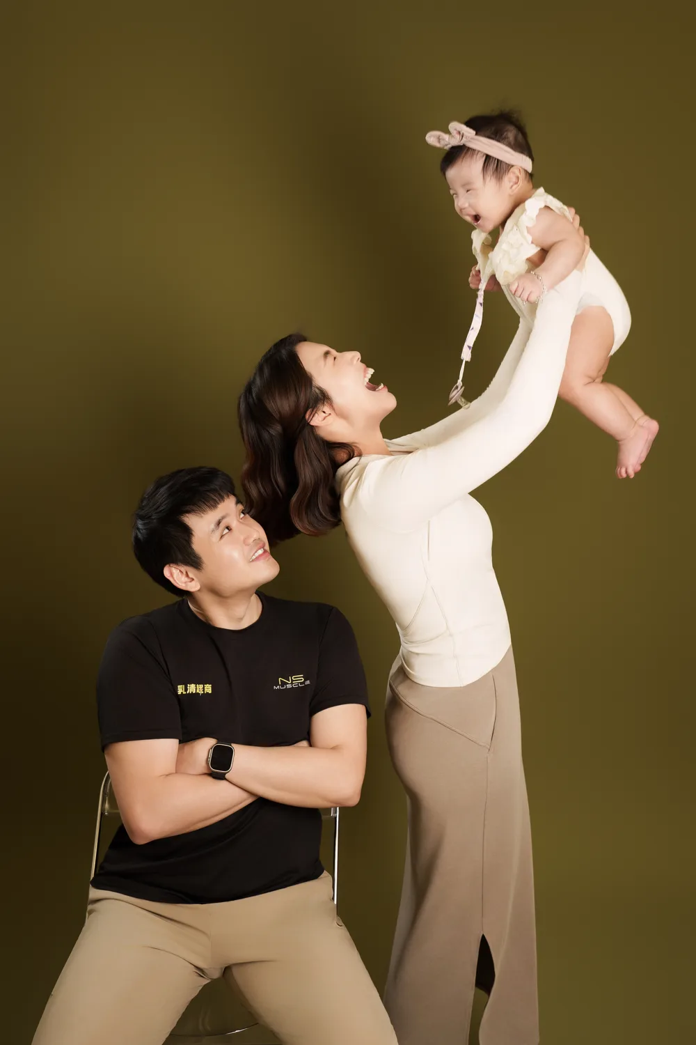 綠色背景親子互動,一位家人舉起孩子,另一位家人在旁看著